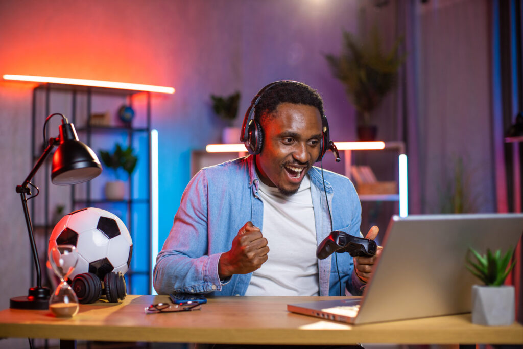 Man wearing headphones sits at a desk with a laptop, gripping a game controller and cheering joyfully in a neon-lit room.