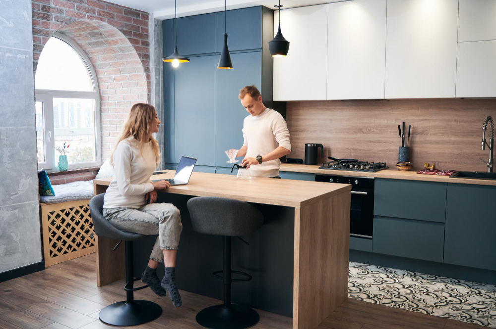 Woman seated at a wooden kitchen island with a laptop, while a man pours coffee beside her in a modern blue-gray kitchen with pendant lights and a brick window wall.