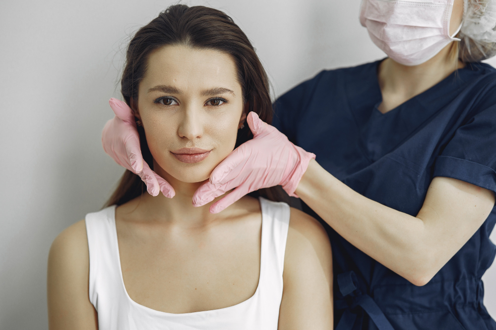 Medical professional in navy scrubs wearing a mask palpating a woman's neck with pink gloves during an examination.