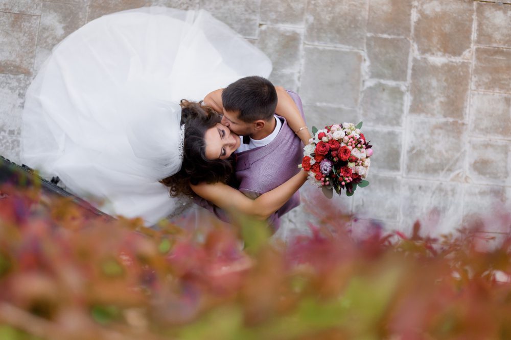 A bride and groom embrace and kiss on a stone courtyard, viewed from above; the bride holds a bouquet of red, pink, and white flowers with greenery in the foreground.