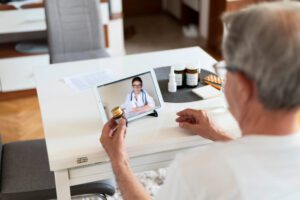 Older man video-calling a doctor on a tablet while holding a pill bottle at a white table with medicines nearby.