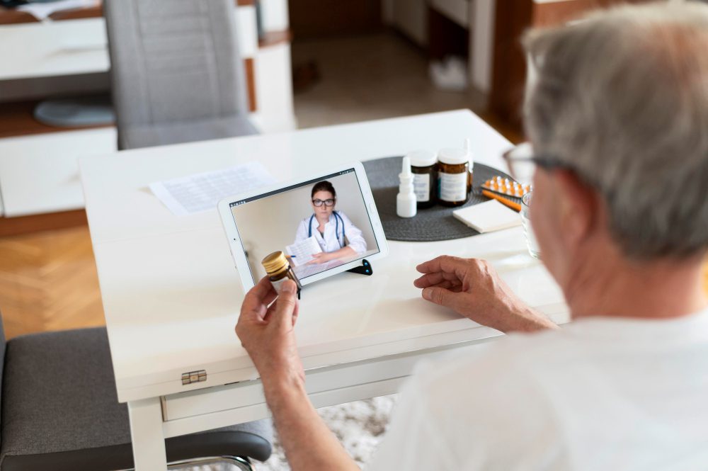 Older man video-calling a doctor on a tablet while holding a pill bottle at a white table with medicines nearby.