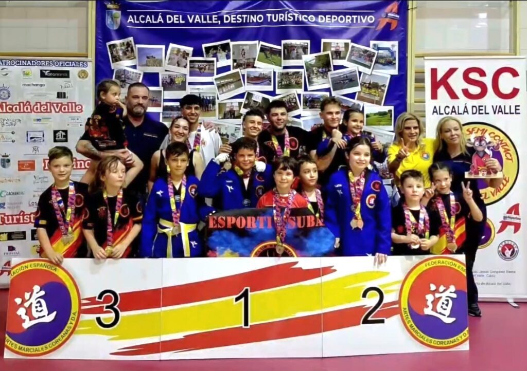 Group of children and coaches on a winners' podium at a martial arts competition, wearing blue and red uniforms with medals, banners reading Alcala del Valle in the background.