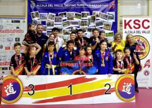 Group of children and coaches on a winners' podium at a martial arts competition, wearing blue and red uniforms with medals, banners reading Alcala del Valle in the background.